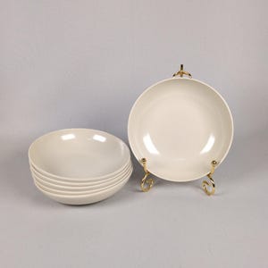 May include: A set of six off-white ceramic bowls with a smooth, glossy finish. Five bowls are stacked neatly on the left, while one bowl is displayed upright on a gold-toned metal wire stand to the right, showcasing the simple, round design.