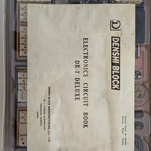 Op de afbeelding: Een vintage elektronica kit met een circuitboek getiteld "Electronics Circuit Book DR-7 Deluxe". De kit bevat diverse elektronische componenten, een Maxell batterij en het "Denshi Block" logo. De kit zit in een doorzichtige plastic container.