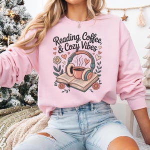 May include: Light pink sweatshirt with the text "Reading, Coffee & Cozy Vibes" and a graphic of headphones, a coffee cup, and an open book. The design includes hearts and flowers. The sweatshirt is a long-sleeved crew neck.