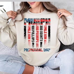 May include: A beige sweatshirt with the word "MURPH" in a patriotic red, white, and blue design. Below the word are the workout details: 1 mile run, 100 pullups, 200 pushups, and 300 squats. The text "MEMORIAL DAY" is at the bottom.