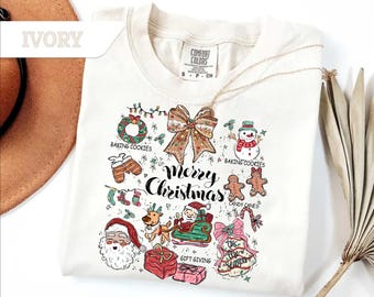 Puede incluir: Camiseta de color marfil con un diseño festivo de "Feliz Navidad". El gráfico incluye un muñeco de nieve, Papá Noel en un trineo, hombres de jengibre y cajas de regalo. La camiseta también presenta un lazo, una corona y texto.