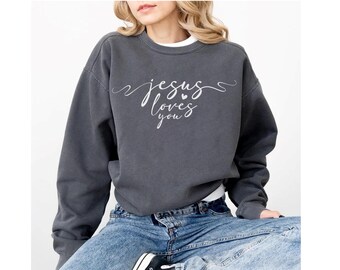 May include: Dark grey sweatshirt with the text "Jesus loves you" in a cursive font. The sweatshirt has a crew neck and long sleeves. The model is wearing blue jeans.