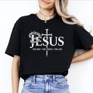May include: Black t-shirt with the word "JESUS" in white, accompanied by a crown of thorns, a cross, and the text "THE WAY &bull; THE TRUTH &bull; THE LIFE". The shirt is made of a soft material.