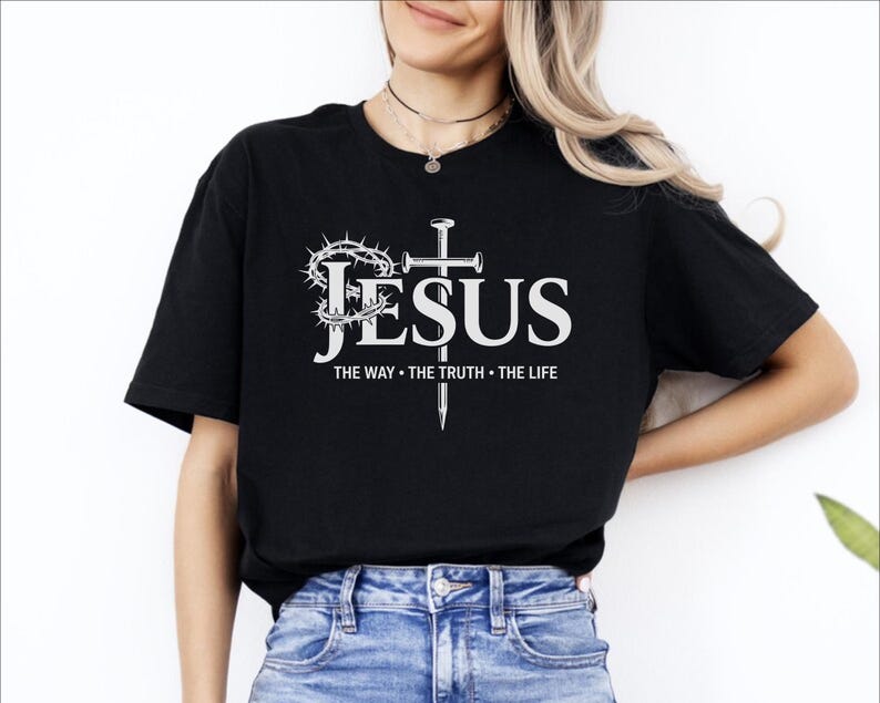 May include: Black t-shirt with the word "JESUS" in white, accompanied by a crown of thorns, a cross, and the text "THE WAY &bull; THE TRUTH &bull; THE LIFE". The shirt is made of a soft material.