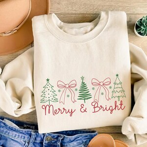 Op de afbeelding: Crèmekleurige sweatshirt met een feestelijk ontwerp. Het heeft groene kerstbomen en rode strikken, met de woorden "Merry & Bright" in rood schrift. De sweatshirt wordt gecombineerd met een blauwe spijkerbroek en een bruine hoed.