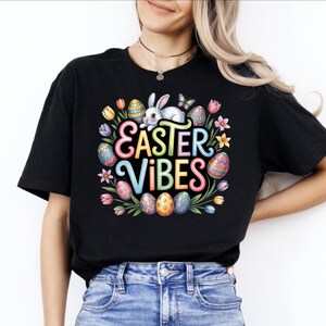 May include: Black t-shirt with a colorful "Easter Vibes" graphic. The design features a white bunny, decorated Easter eggs, flowers, and the text "Easter Vibes" in a rainbow font. The shirt is made of a soft material.