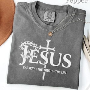 May include: A gray t-shirt with the word "JESUS" in white, along with a crown of thorns and a cross. The text "THE WAY &bull; THE TRUTH &bull; THE LIFE" is below. A gold necklace is draped on the shirt.
