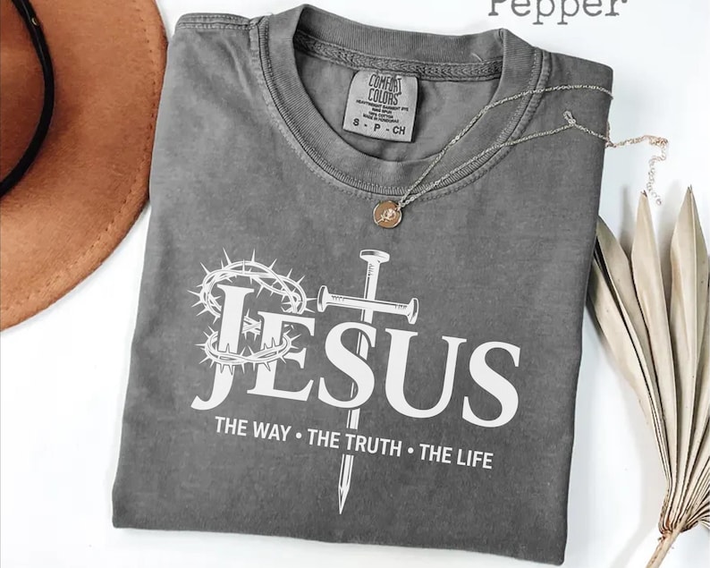 May include: A gray t-shirt with the word "JESUS" in white, along with a crown of thorns and a cross. The text "THE WAY &bull; THE TRUTH &bull; THE LIFE" is below. A gold necklace is draped on the shirt.