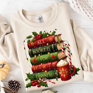 May include: Cream-colored sweatshirt featuring a stack of books with the words "ALL BOOKED FOR CHRISTMAS." The books are red and green, decorated with holly and string lights. A red mug of hot cocoa and candy canes complete the festive design.