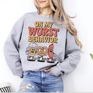 May include: A light grey sweatshirt featuring the phrase "ON MY WURST BEHAVIOR" in orange and yellow. The design includes cartoon images of a beer stein, pretzel, and sausage. A casual, comfortable style.