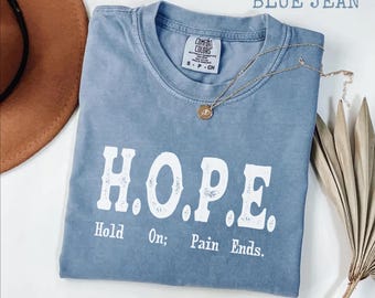 May include: A blue jean Comfort Colors t-shirt with the word "H.O.P.E." printed in white. The text "Hold On; Pain Ends." is below the main word. A gold necklace with a pendant is on the shirt.