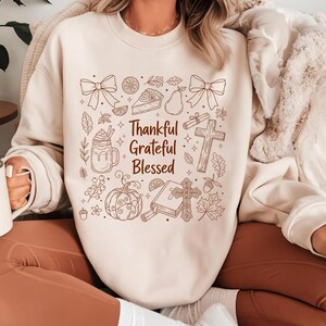 May include: Cream-colored sweatshirt with a brown graphic design featuring bows, a slice of pie, a pear, a cross, and the words "Thankful Grateful Blessed." The design has a fall or Thanksgiving theme.