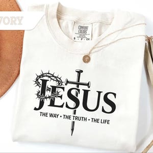 May include: Ivory-colored t-shirt with the word "JESUS" in bold black letters, a crown of thorns, a cross, and the text "THE WAY &bull; THE TRUTH &bull; THE LIFE." A gold necklace is draped over the shirt.