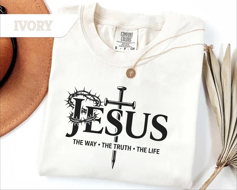 May include: Ivory-colored t-shirt with the word "JESUS" in bold black letters, a crown of thorns, a cross, and the text "THE WAY &bull; THE TRUTH &bull; THE LIFE." A gold necklace is draped over the shirt.