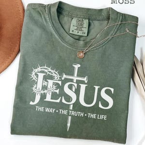 May include: A moss green t-shirt with the word "JESUS" in white, along with a crown of thorns, a cross, and the text "THE WAY &bull; THE TRUTH &bull; THE LIFE." A gold necklace with a pendant is draped across the shirt.