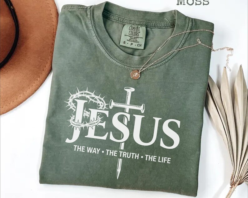 May include: A moss green t-shirt with the word "JESUS" in white, along with a crown of thorns, a cross, and the text "THE WAY &bull; THE TRUTH &bull; THE LIFE." A gold necklace with a pendant is draped across the shirt.