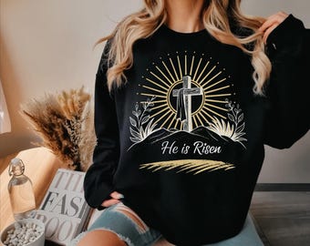 May include: Black sweatshirt with a graphic of a cross on a hill, surrounded by a sunburst design and foliage. The text "He is Risen" is printed below the cross in a script font. The sweatshirt is a long-sleeved crew neck.