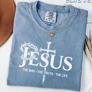 May include: A blue jean Comfort Colors t-shirt with the word "JESUS" in white, along with a cross, crown of thorns, and the text "THE WAY &bull; THE TRUTH &bull; THE LIFE." A gold necklace and a brown hat are also visible.