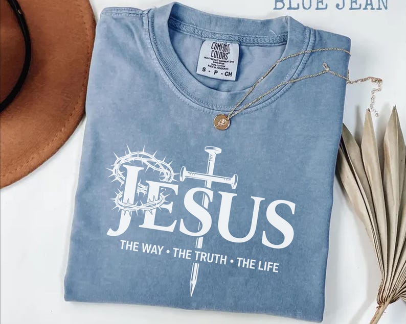 May include: A blue jean Comfort Colors t-shirt with the word "JESUS" in white, along with a cross, crown of thorns, and the text "THE WAY &bull; THE TRUTH &bull; THE LIFE." A gold necklace and a brown hat are also visible.