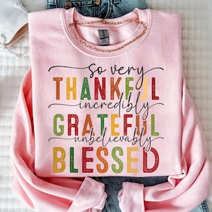 May include: A light pink sweatshirt with the words "So very THANKFUL, incredibly GRATEFUL, unbelievably BLESSED" in colorful lettering. A gold chain necklace is draped on the sweatshirt. White sneakers and jeans are visible.