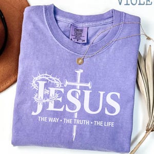 May include: A violet Comfort Colors t-shirt with the word "JESUS" in white, along with a cross and crown of thorns graphic. The text "THE WAY &bull; THE TRUTH &bull; THE LIFE" is below. A gold necklace and a brown hat are also visible.