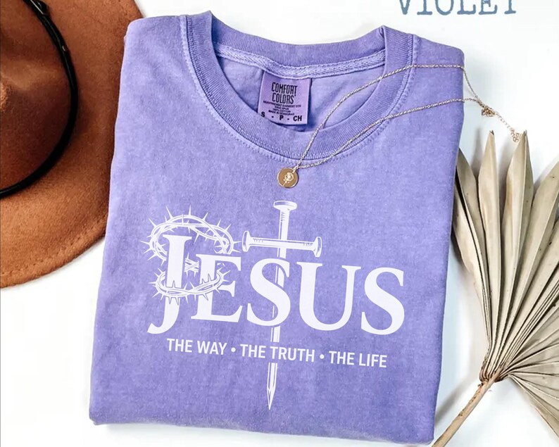 May include: A violet Comfort Colors t-shirt with the word "JESUS" in white, along with a cross and crown of thorns graphic. The text "THE WAY &bull; THE TRUTH &bull; THE LIFE" is below. A gold necklace and a brown hat are also visible.