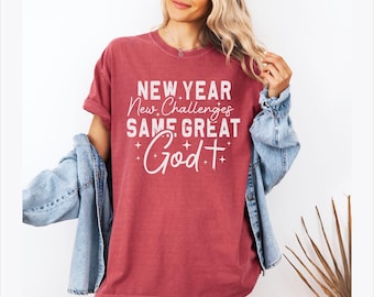 May include: A red t-shirt with the text "NEW YEAR New Challenges SAME GREAT God" in white script. The shirt has a crew neck and short sleeves. A light blue denim jacket is draped over the shoulders.