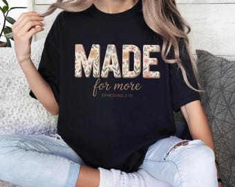 May include: Black t-shirt with the words "MADE for more" in a floral design. The text "Ephesians 2:10" is below the main text. The shirt is worn by a person.