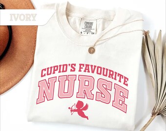 以下が含まれることがあります： アイボリー色のTシャツ。ピンク色の文字で「Cupid's Favourite Nurse」と書かれています。テキストの下には、弓矢を持ったキューピッドの小さなグラフィックがあります。金のネックレスがシャツの上にかけられています。