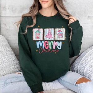 Op de afbeelding: Donkergroene sweatshirt met de tekst "Merry Christmas" in kleurrijke tekst. De sweatshirt heeft drie rechthoekige ontwerpen met een huis, een kerstboom en een ornament. Het ontwerp is feestelijk en geschikt voor de feestdagen.