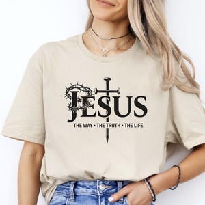 May include: Beige t-shirt with the word "JESUS" in large black letters, a cross, and a crown of thorns. Below the word "JESUS" are the words "THE WAY &bull; THE TRUTH &bull; THE LIFE" in black. The shirt is a casual, everyday wear item.