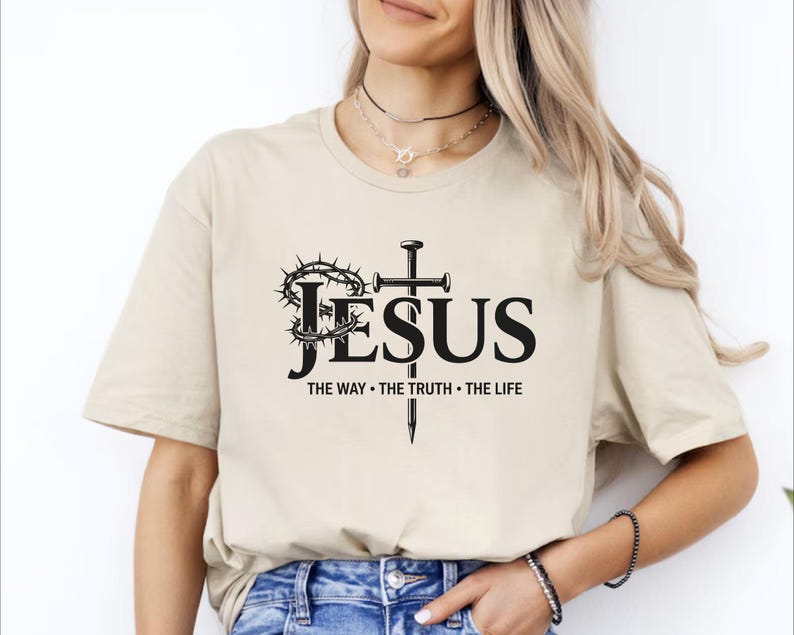 May include: Beige t-shirt with the word "JESUS" in large black letters, a cross, and a crown of thorns. Below the word "JESUS" are the words "THE WAY &bull; THE TRUTH &bull; THE LIFE" in black. The shirt is a casual, everyday wear item.