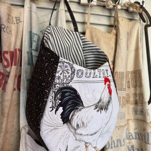 May include: A handmade tote bag with a rooster design. The bag features a black and white rooster illustration on a white background, with a black and white striped interior and a brown star patterned exterior. The bag is hanging on a white wooden shutter.