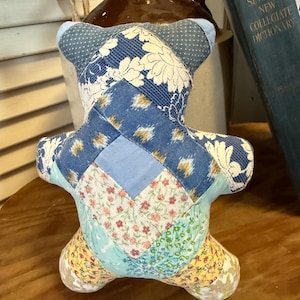 May include: A handmade patchwork teddy bear in shades of blue, white, and yellow. The bear is constructed from various fabric patterns, including floral and geometric designs. The bear has a vintage, handcrafted aesthetic.
