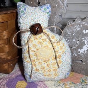May include: A stuffed cat figure crafted from quilted fabric featuring a patchwork of floral patterns in yellow, blue, and white. The cat is adorned with a brown metal jingle bell tied around its neck with a piece of natural jute twine.