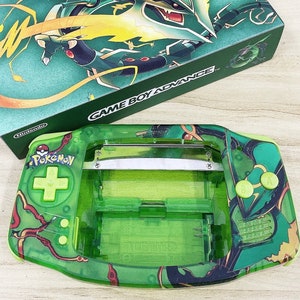 May include: A green Game Boy Advance console and matching box. The console features a transparent green shell with a Pokemon design. The box and console have a green and gold color scheme with the text "GAME BOY ADVANCE" and "Pokemon".
