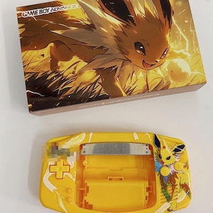 May include: A yellow Game Boy Advance SP console with a custom design featuring a yellow Eevee with a white and blue lightning bolt. The console is open and the screen is visible. The console is on a white surface. The text "GAME BOY ADVANCE" is visible on the box.