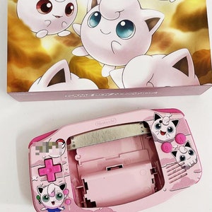 May include: A pink Game Boy Advance SP console with a Jigglypuff design. The console is open and the screen is visible. The console is on a white surface. The text "(Nintendo)" is visible on the console.
