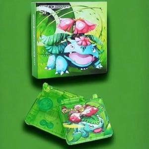 May include: A green Game Boy Advance SP console with a clear green shell. The console features a Pokemon image of Venusaur on the front. The image shows Venusaur with a pink flower on its head and green leaves.