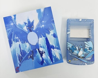 May include: A blue Game Boy Color with a blue and white Articuno Pokemon design. The Game Boy Color is next to a blue box with a blue Articuno Pokemon design.