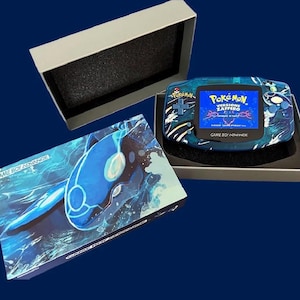 Op de afbeelding: Een doorschijnende blauwe Game Boy Advance handheld console met een Kyogre-afbeelding, gepresenteerd in een grijze doos met zwarte schuiminzetstukken. Het scherm toont de titel Pokémon Versione Zaffiro. Een bijpassend deksel met Kyogre-kunst is inbegrepen.