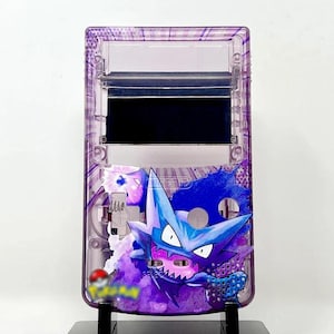 May include: A purple and blue Game Boy Color with a custom design featuring a Pokemon character. The character is a blue and white creature with sharp teeth and a purple background. The Game Boy Color is on a black stand.