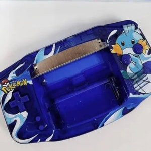 May include: A blue Game Boy Advance SP with a Pokemon design. The design features a blue and white wave pattern with a cartoon character of a blue and white Pokemon with a yellow fin. The text "Pokemon" is printed on the front of the device.