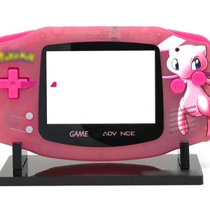 May include: Pink Game Boy Advance SP with a pink and white Jigglypuff design on the back. The console is on a black stand. The screen is blank and the buttons are visible. The text "GAME ADVANCE" is printed on the front of the console.