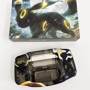 May include: A clear plastic Game Boy Advance SP with a black and white design featuring a Pokemon character. The design includes a black and white image of a Pokemon character with yellow accents. The Game Boy Advance SP is a handheld video game console.