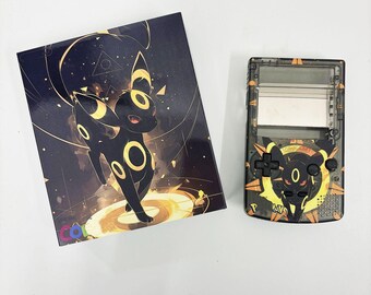 May include: A black and gold Game Boy Color with a custom skin featuring a black and yellow Pokemon character. The character has large, round eyes and a yellow ring around its neck.