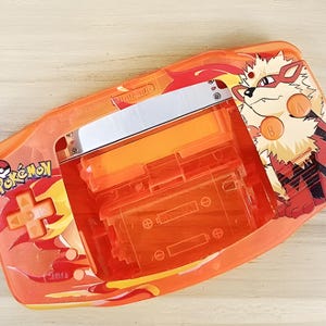 May include: A clear orange Game Boy Advance SP console with a Pokemon Fire Red design. The console features a graphic of the Pokemon character, Flareon, and the text "Pokemon" on the front.