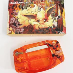 May include: Orange Game Boy Advance shell with a custom design featuring a fiery Eevee Pokemon. The shell has a clear plastic front and a black and white button.