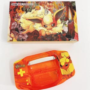 May include: Orange Game Boy Advance console with a clear shell and a custom design featuring a cartoon image of a Vaporeon Pokemon. The console has a yellow button and a yellow and orange design.