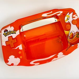 May include: Orange and white handheld video game console with a Pokemon logo and a white character on the screen. The console has a slot for a game cartridge and a screen.